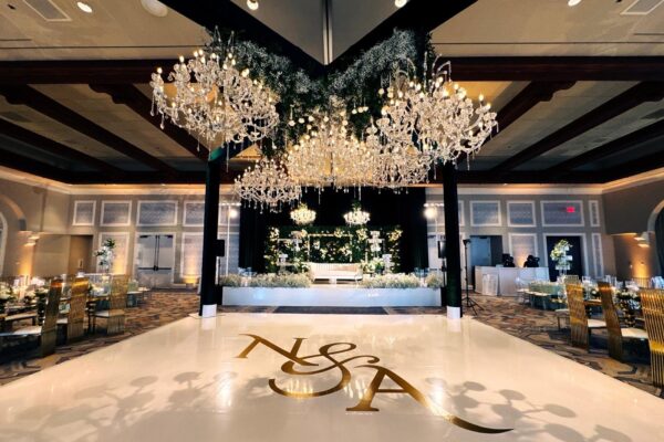 Black box truss with crystal chandeliers and greenery over a white dance floor with a gold N&A monogram