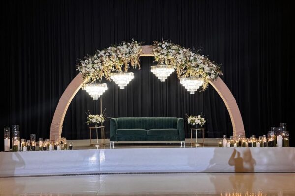 Fabric-wrapped arch with white florals and crystal chandeliers over a green velvet sofa on a white stage