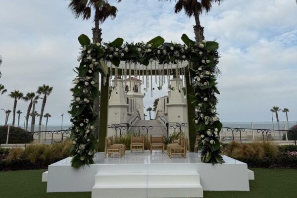 Outdoor mandap wrapped in white roses and greenery on a white platform with gold ceremony chairs