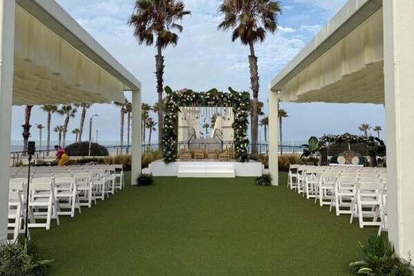 Outdoor floral mandap at the end of a ceremony aisle lined with white chairs and draped cabanas